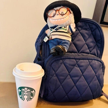 Plush toy with glasses sitting on a blue backpack next to a Starbucks cup on a table.
