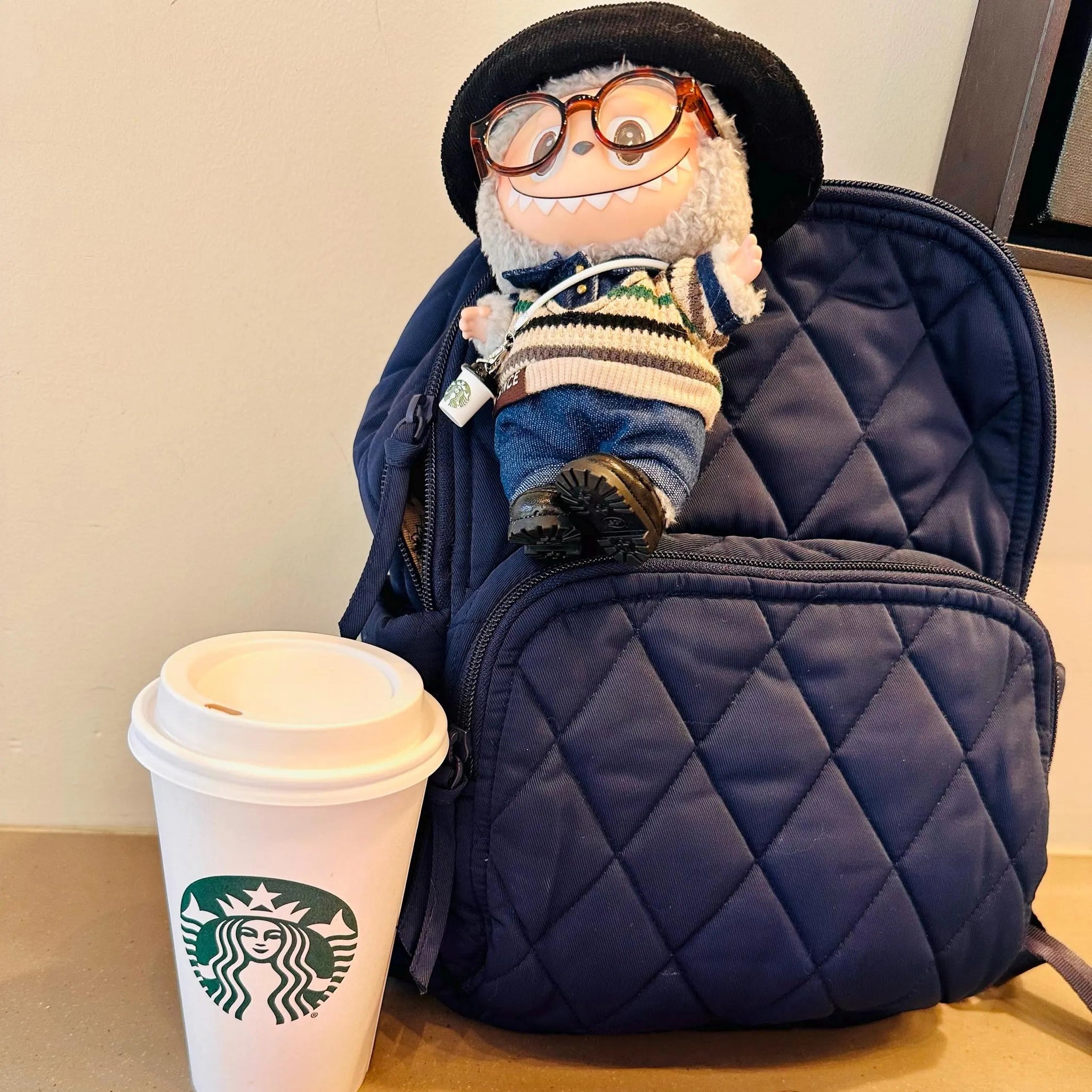 Plush toy with glasses sitting on a blue backpack next to a Starbucks cup on a table.